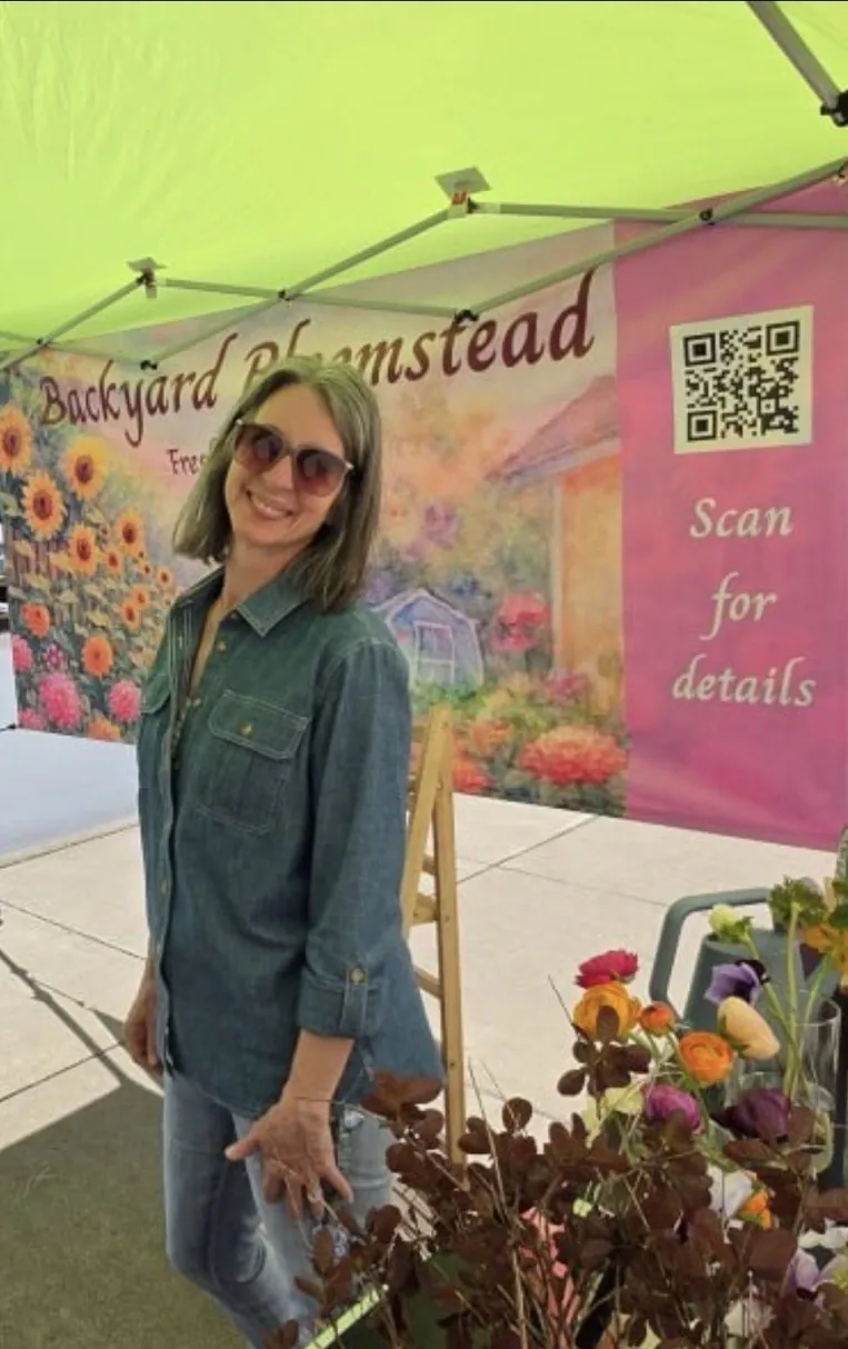 Susette smiling at the Backyard Bloomstead market booth, with a painted flower banner and a bucket of fresh-cut flowers