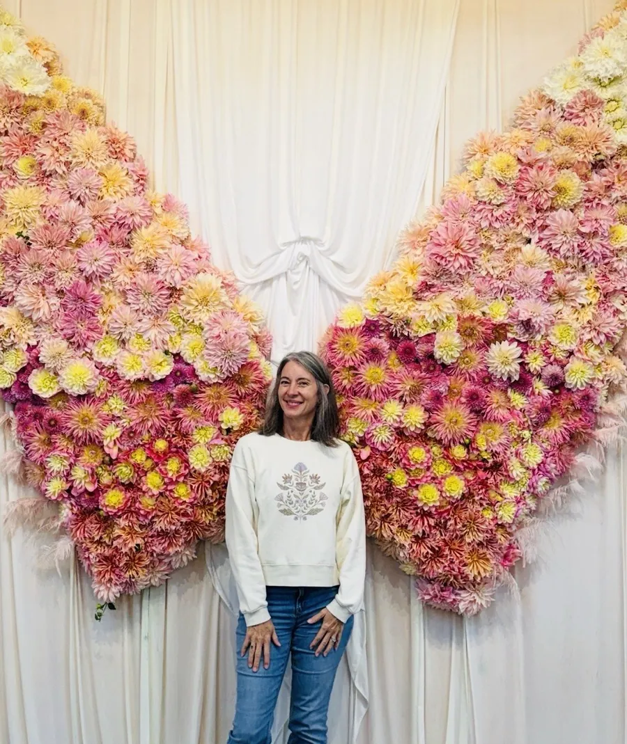 Susette standing in front of a pair of giant wings made of pink, peach, and yellow dahlias